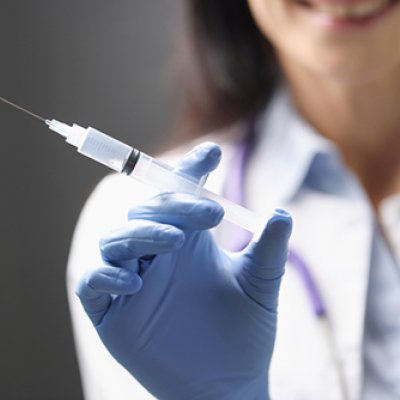 Close up of gloved woman's hand holding a syringe, with her trunk blurred in the background.